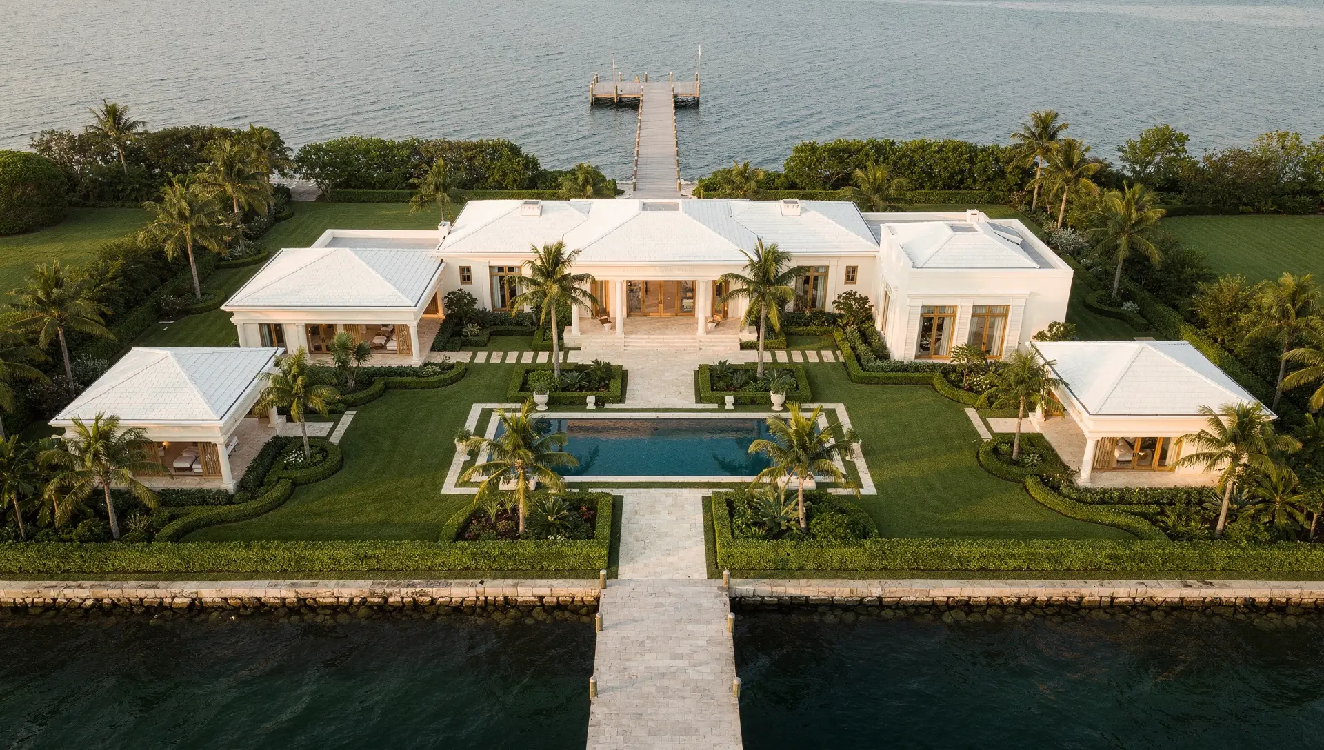An aerial view of a white waterfront estate with a formal pool, gardens and a private dock