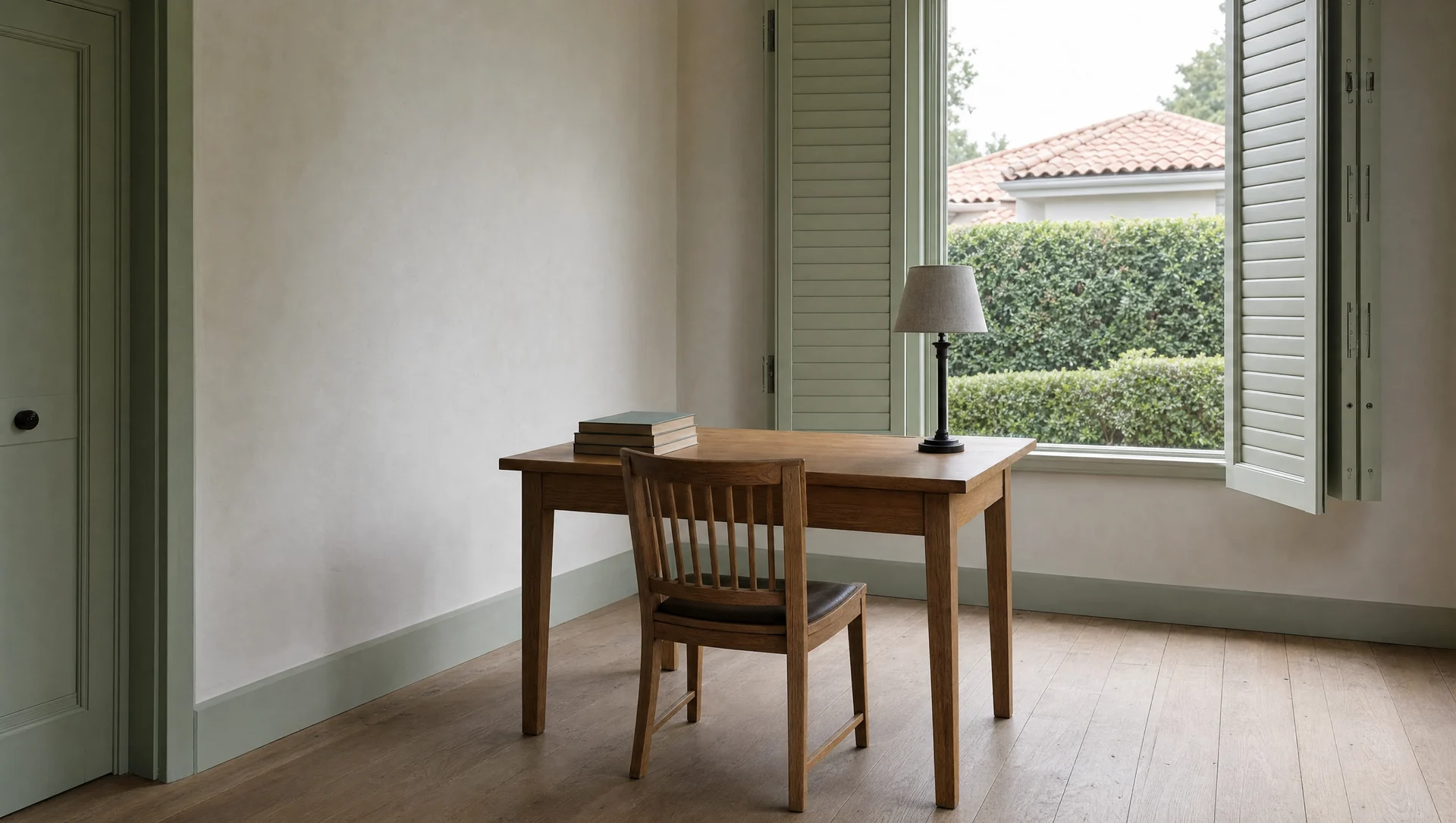 A plain writing table and chair at a shuttered window, with a hedge and a tiled roof beyond.